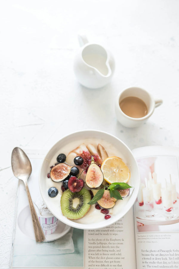 Breakfast bowl with figs, kiwi, blueberries and lemon on a white table next to a coffee cup and an open book, photo by Brooke Lark