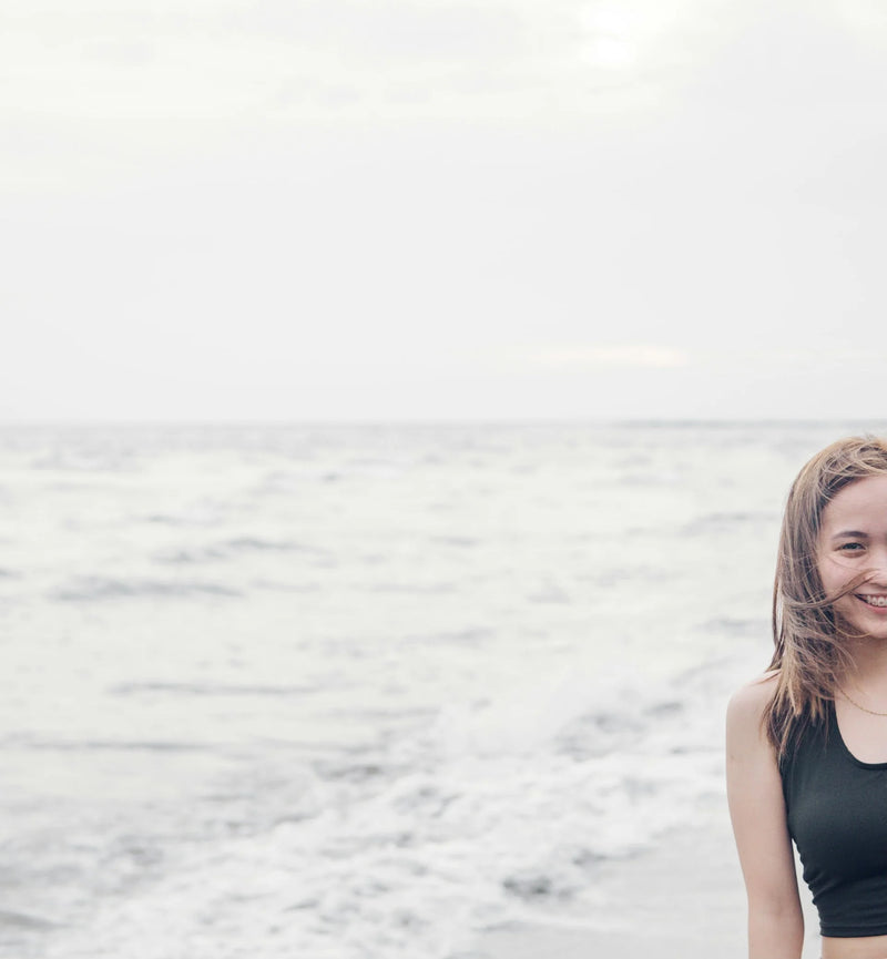 Smilende kvinde på en strand foran et roligt gråt hav, med vinden i håret
