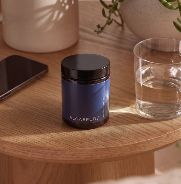 PersonaPath AlgaePure supplement jar on a wooden side table, beside a glass of water and a phone