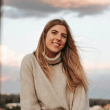 Smiling woman with long hair in a beige knit sweater sitting outdoors at dusk, photo by Andrea Ba