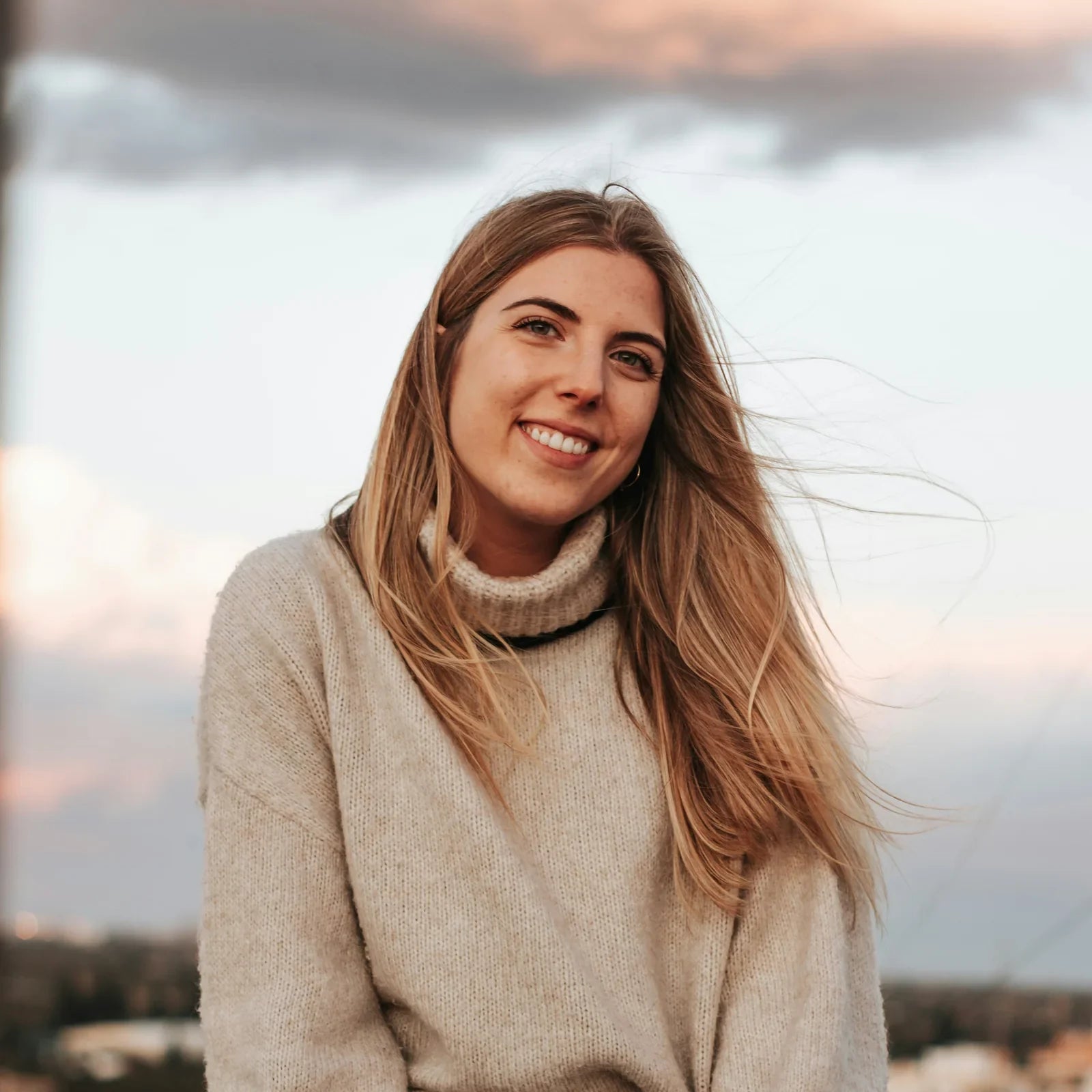 Smiling woman with long hair in a beige knit sweater sitting outdoors at dusk, photo by Andrea Ba