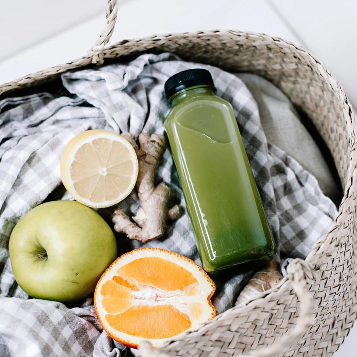 Kurv med en flaske grøn juice, et æble, en citron, en halv appelsin og ingefær på et ternet stykke stof, foto af Birgith Roosipuu