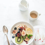 Morgenmadsskål med figner, kiwi, blåbær og citron på et hvidt bord ved siden af en kaffekop og en åben bog, foto af Brooke Lark