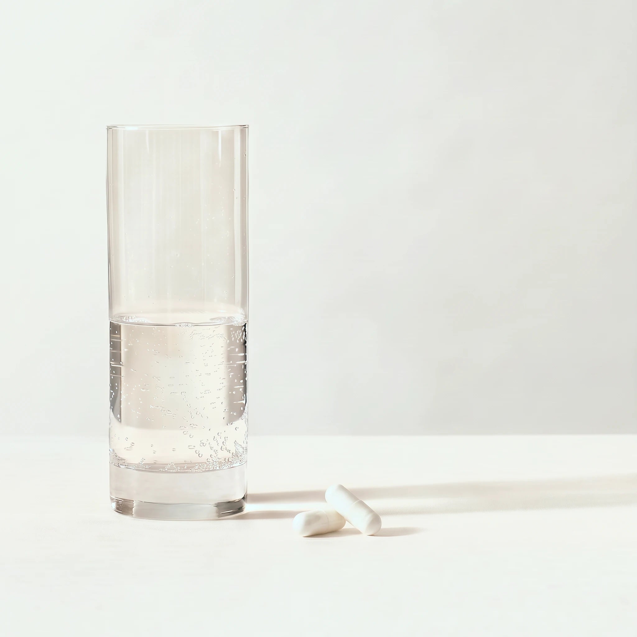 Højt glas halvt fyldt med brusende vand ved siden af to hvide kosttilskudskapsler på en blød lys gråbeige baggrund