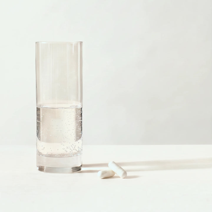 Højt glas halvt fyldt med brusende vand ved siden af to hvide kosttilskudskapsler på en blød lys gråbeige baggrund