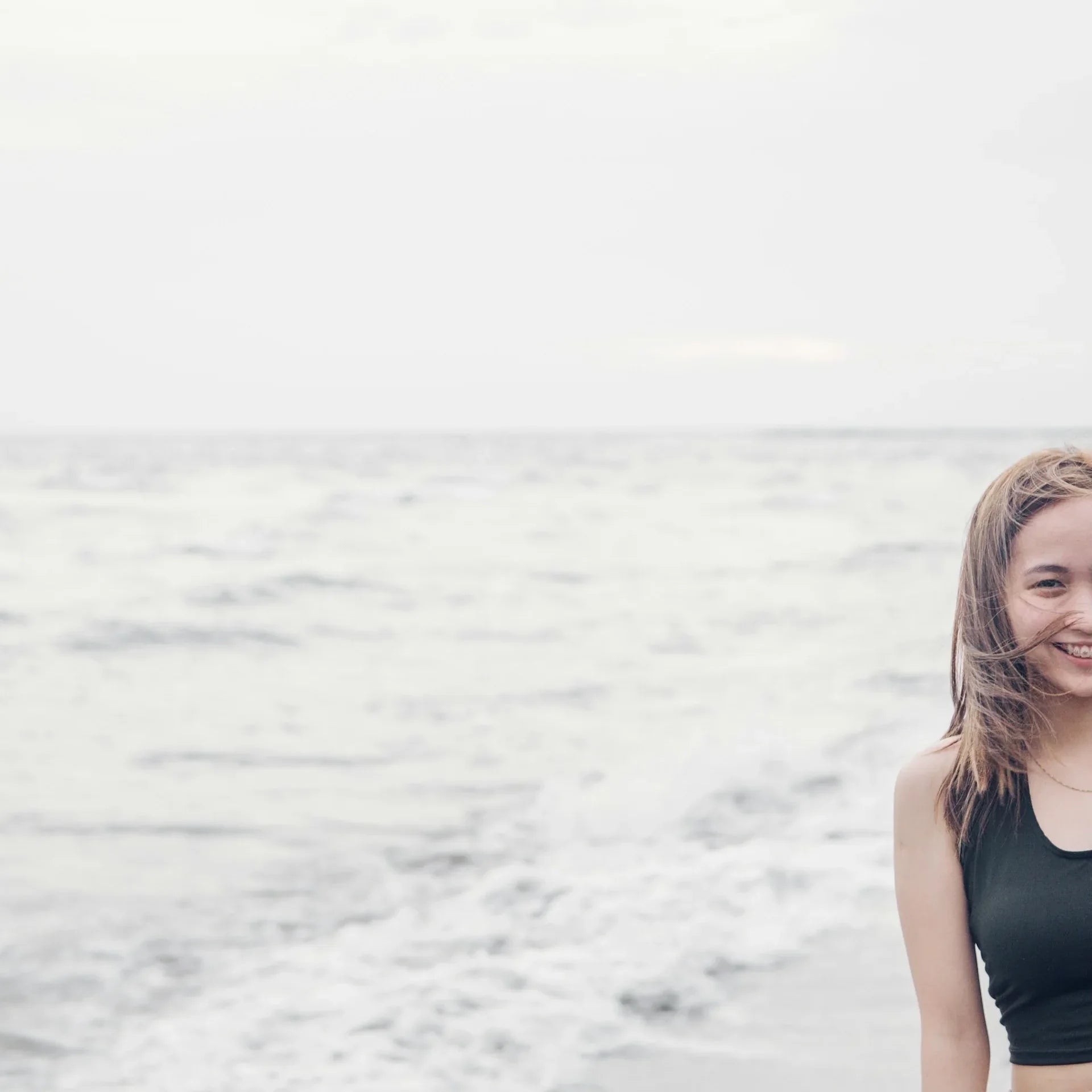 Smilende kvinde på en strand foran et roligt gråt hav, med vinden i håret