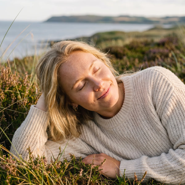 En kvinde hviler i lyng ved havet i gyldent lys, med lukkede øjne og et smil