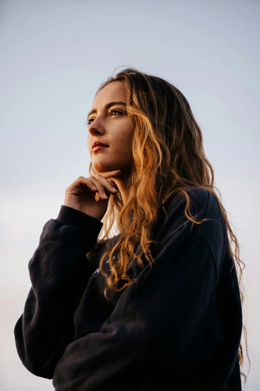 Woman with long wavy hair in a dark sweatshirt resting her chin on her hand, looking away against a pale sky, photo by Raphael Nast
