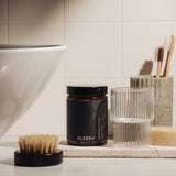 PersonaPath Sleep+ supplement jar on a stone tray in a bathroom, beside a ribbed glass and a bamboo toothbrush holder
