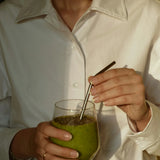 Person in a white shirt holding a glass of green smoothie with a metal straw, photo by Victoria Aleksandrova